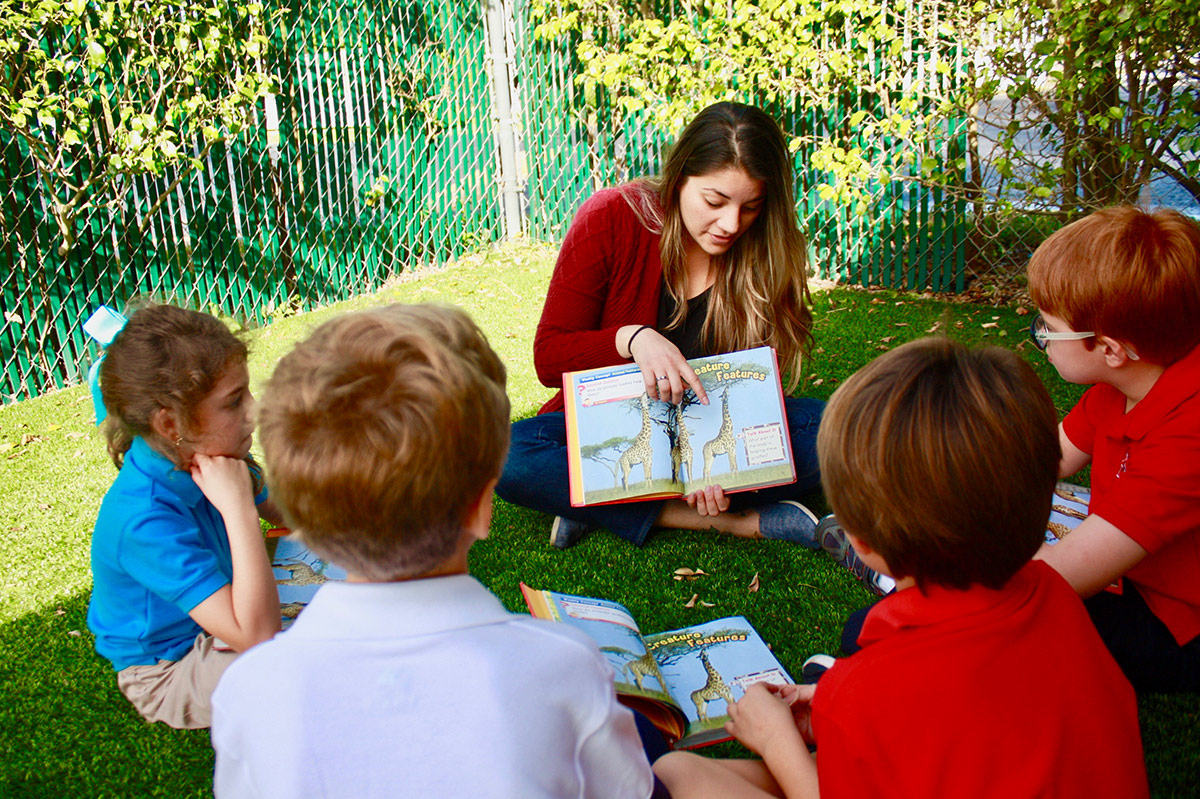 A teacher reading a book to children outside while sitting on the grass