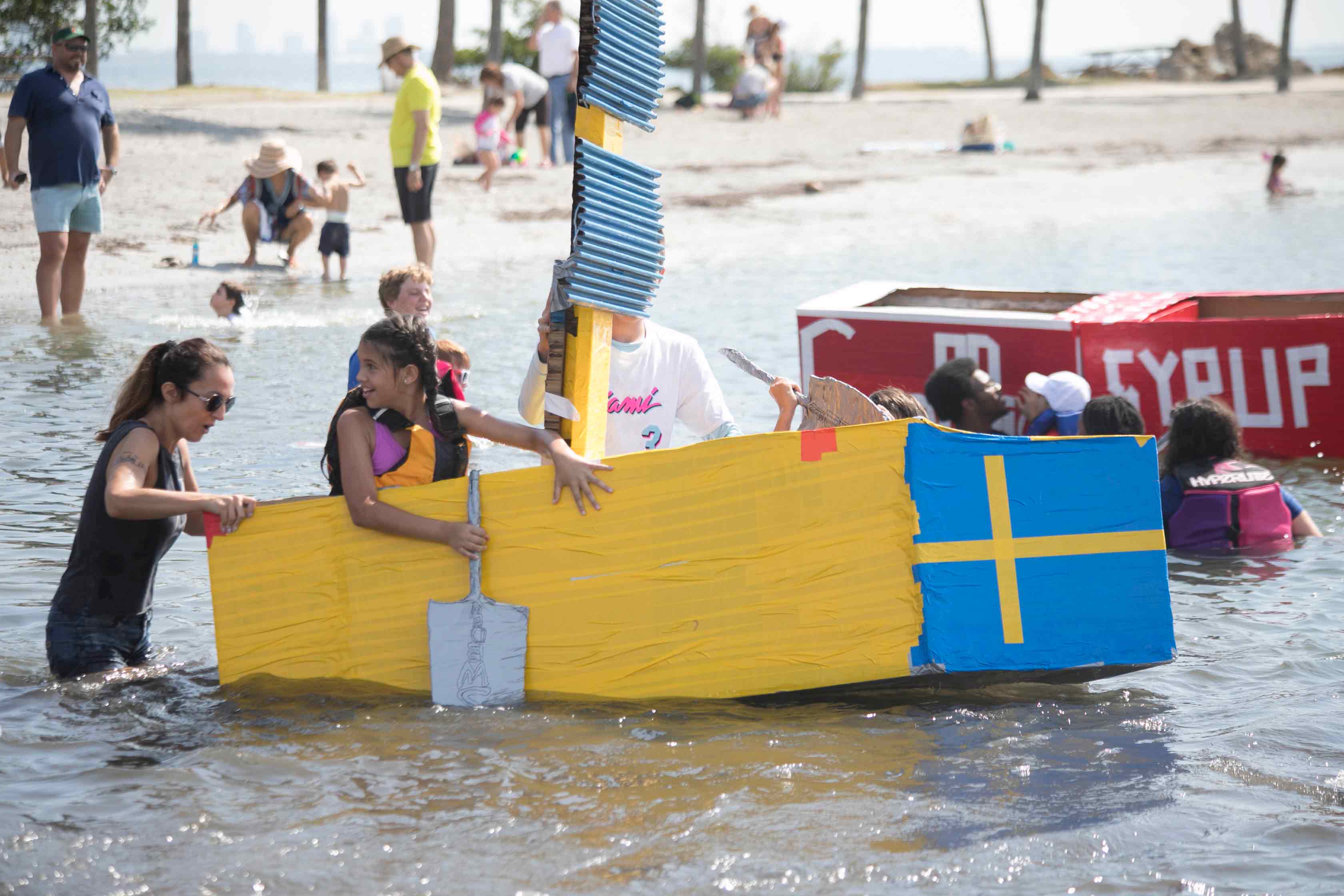 girl rowing a cardboard boat with parent behind her