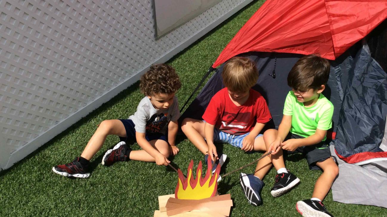 3 kids sitting outside a tent roasting marshmallows on a pretend fire made of paper