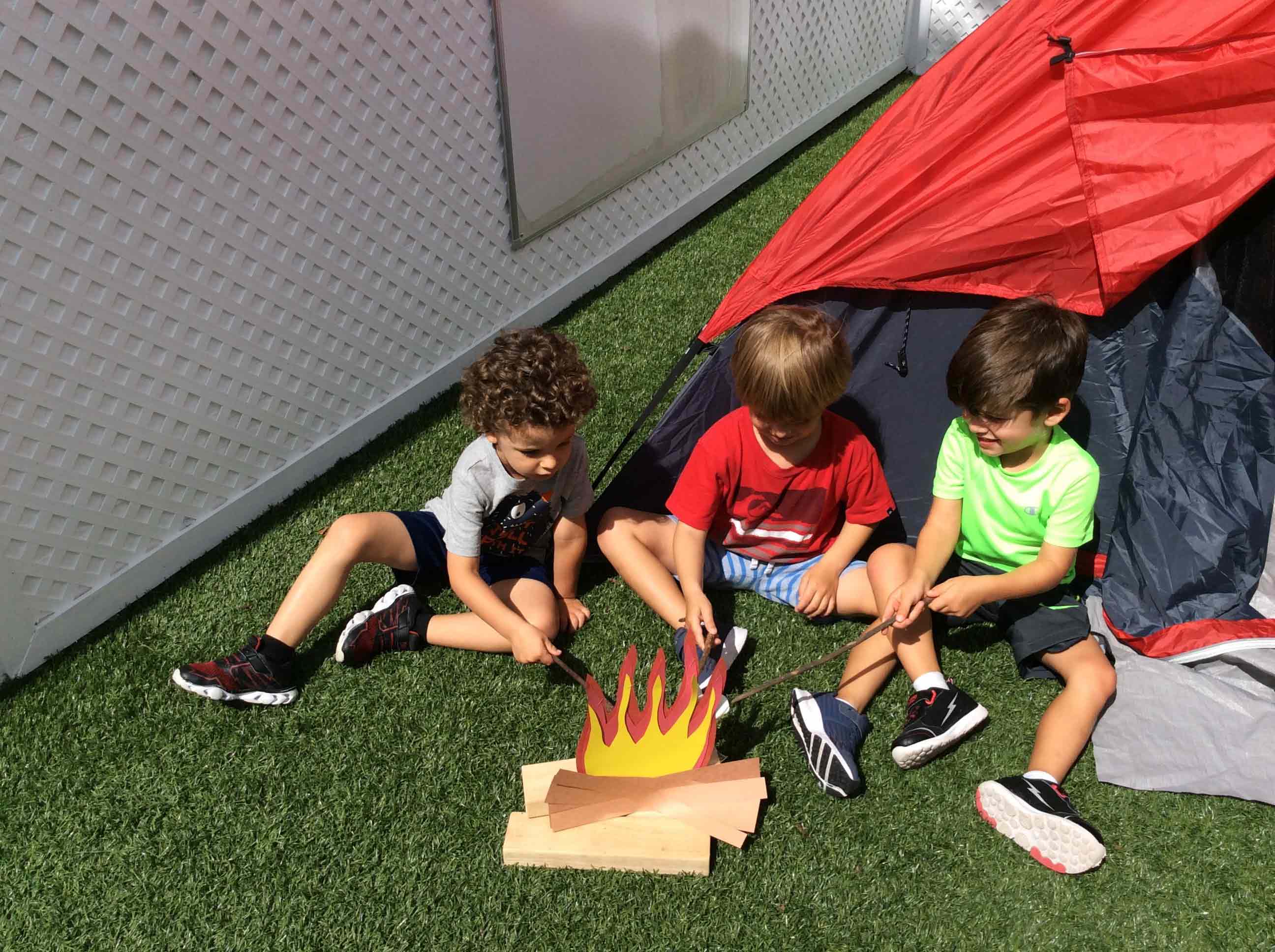 3 kids sitting outside a tent roasting marshmallows on a pretend fire made of paper